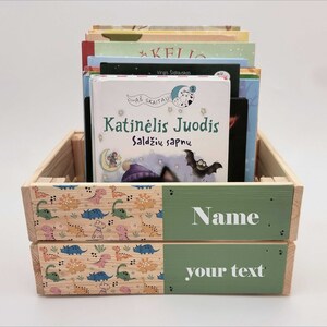 May include: A wooden crate with a green and dinosaur print design. The crate is filled with children's books. The front of the crate has a green panel with the text "Name your text".