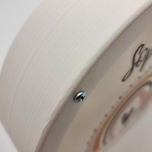May include: A white wooden round sign with a clear plastic overlay. The overlay features a rainbow design with the name "Sophia's" written in black cursive.