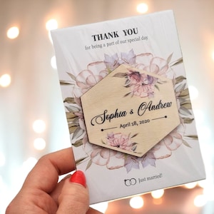May include: A wedding favour card with floral details and a wooden hexagon. The card says "THANK YOU" and "Sophia & Andrew, April 18, 2020." The words "just married!" and two wedding rings are at the bottom.