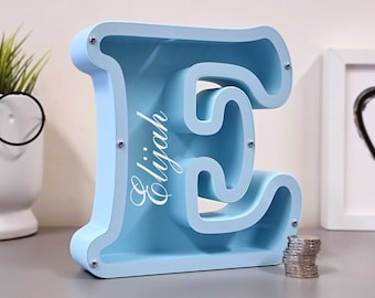 May include: A light blue wooden piggy bank shaped like the letter E. The name "Elijah" is written on the front of the bank in white lettering. A stack of coins is sitting in front of the bank.