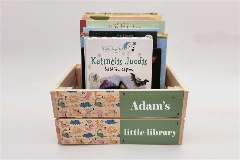 May include: A wooden crate with a green and white label that says "Adam's little library". The crate is filled with children's books, including one with the title "Katinėlis Juodis Saldžių Sapnų".