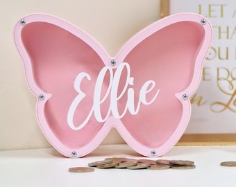 May include: Pink butterfly-shaped wooden piggy bank with the name "Ellie" written in white on the front.