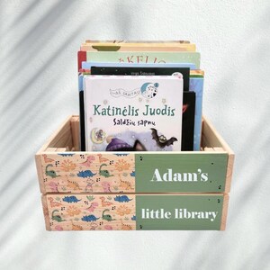 May include: A wooden crate with a green and white label that says "Adam's little library". The crate is filled with children's books, including one with the title "Katinėlis Juodis Saldžių sapnų".