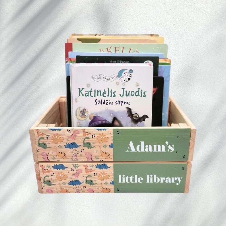 May include: A wooden crate with a green and white label that says "Adam's little library". The crate is filled with children's books, including one with the title "Katinėlis Juodis Saldžių sapnų".