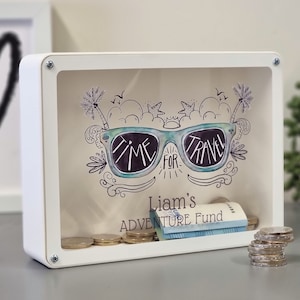 May include: White shadow box with a clear front, featuring a hand-drawn design of sunglasses with the words "TIME FOR TRAVEL." The box is labeled "Liam's ADVENTURE FUND" and contains coins and folded bills.