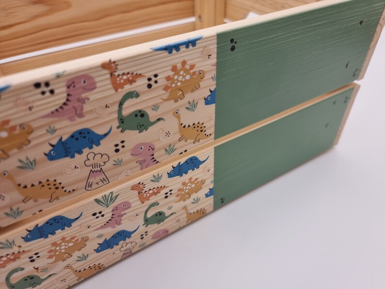 May include: A wooden crate with a green painted top and a dinosaur pattern on the sides. The crate is made of natural wood and has a rustic look.