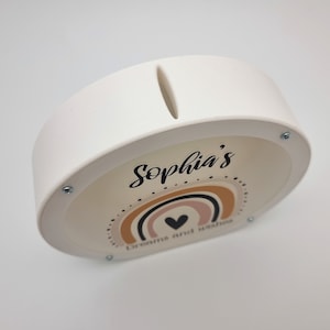May include: White ceramic piggy bank with a slot on top. The front features a rainbow design with a heart and the text "Sophia's Dreams and Wishes".