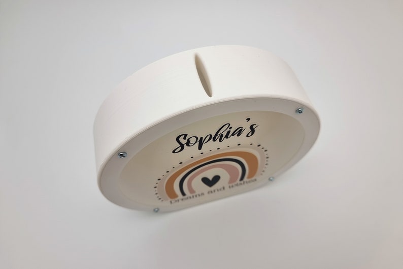 May include: White ceramic piggy bank with a slot on top. The front features a rainbow design with a heart and the text "Sophia's Dreams and Wishes".