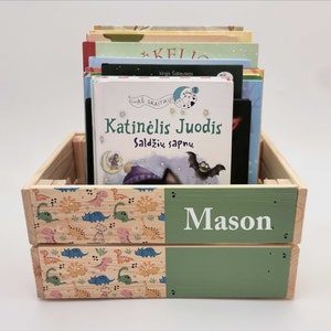 May include: A wooden crate with a green and dinosaur-patterned design. The crate is filled with books, including one titled "Katinėlis Juodis Saldžių sapnu". The crate is personalised with the name "Mason" on the front.