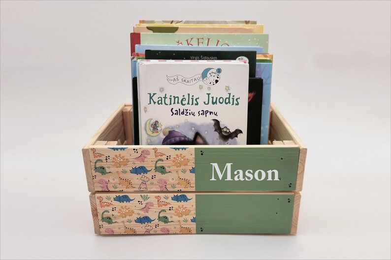May include: A wooden crate with a green and dinosaur-patterned design. The crate is filled with books, including one titled "Katinėlis Juodis Saldžių sapnu". The crate is personalised with the name "Mason" on the front.