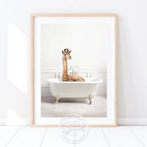 Könnte beinhalten: Gerahmter Druck, der eine Giraffe in einer weißen Badewanne mit Löwenfüßen zeigt. Die Giraffe, mit ihren braunen und beigen Flecken, befindet sich in der Wanne mit silbernen Wasserhähnen. Der Druck ist vor einer weißen Wand platziert und erzeugt eine skurrile Szene.