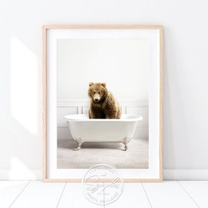 May include: A framed print featuring a brown bear sitting in a white clawfoot bathtub. The bear is centered in the tub, with a light background. The print is in a light wood frame. The image has a whimsical, yet elegant feel.