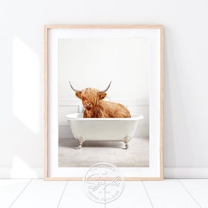 May include: A framed print featuring a Highland cow relaxing in a white clawfoot bathtub. The cow has long, shaggy, reddish-brown fur and large horns. The bathroom setting is minimalist with a light wood frame.