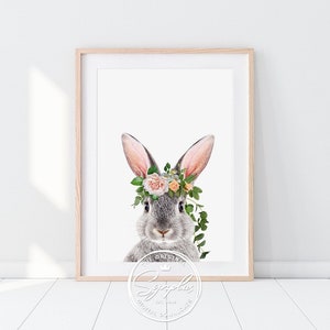 Puede incluir: Una impresión enmarcada con un conejo gris adornado con una corona floral de rosas rosas y melocotón, hojas verdes y vides. El conejo está centrado sobre un fondo blanco. La impresión está en un marco de madera clara. Un logotipo circular blanco está en la parte inferior.