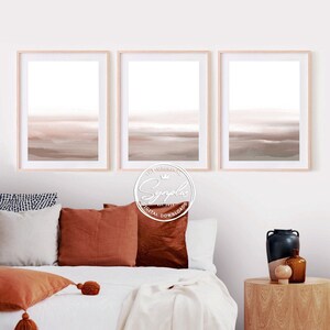 Könnte beinhalten: Drei gerahmte abstrakte Aquarelldrucke in gedeckten Farben in Beige, Braun und Grau. Die Drucke hängen an einer weißen Wand über einem Bett mit weißen Bettwäsche und orangefarbenen Kissen.