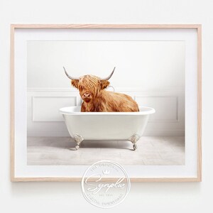 Könnte beinhalten: Eine Highland-Rinderkuh, die ein Bad in einer weißen Klauenfußbadewanne nimmt. Die Kuh ist braun und hat langes, zotteliges Fell. Die Badewanne steht auf einem gefliesten Boden. Das Bild ist in einem hellen Holzrahmen gerahmt.