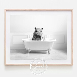 Könnte beinhalten: Schwarz-Weiß-Druck in einem Rahmen mit einem Nilpferd in einer Badewanne mit Löwenfüßen. Das Nilpferd befindet sich im Zentrum der Wanne. Der Hintergrund ist minimalistisch, mit weißen Wänden und einem gefliesten Boden. Der Druck ist in einem hellen Holzrahmen.