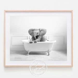 May include: Black and white framed print featuring a baby elephant sitting in a claw-foot bathtub. The elephant is the focal point, with its large ears and trunk visible. The background is a minimalist white bathroom setting.
