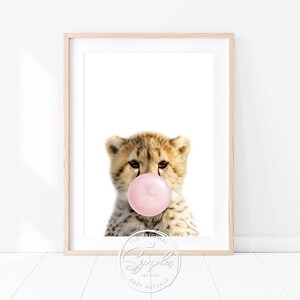 Könnte beinhalten: Ein süßes Gepardenjunges mit einem rosa Kaugummi im Maul. Der Gepard schaut direkt in die Kamera. Der Hintergrund ist weiß.