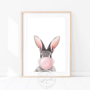 Könnte beinhalten: Ein graues Kaninchen mit rosa Ohren bläst eine rosa Blase. Das Kaninchen schaut direkt in die Kamera. Das Bild ist in einem weißen Rahmen.