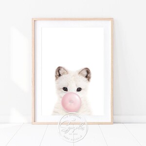 Könnte beinhalten: Ein weißer Polarfuchs-Welpe mit rosa Kaugummi im Maul. Der Fuchs schaut direkt auf den Betrachter. Das Bild ist in einem weißen Rahmen mit einer hellbraunen Umrandung.
