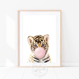 Könnte beinhalten: Ein süßes Tigerjunges mit blauen Augen bläst eine rosa Blase. Der Tiger sitzt auf einem weißen Hintergrund. Das Bild ist in einem hellen Holzrahmen gerahmt.