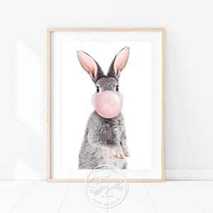 Könnte beinhalten: Ein graues Kaninchen mit rosa Ohren bläst eine rosa Blase. Das Kaninchen schaut direkt in die Kamera. Der Hintergrund ist weiß. Das Bild enthält den Text "The Original" und "Cuddles" mit einem Logo eines Kaninchens mit einem Herzen.