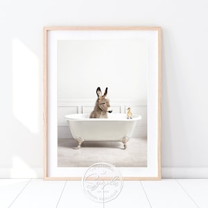 Könnte beinhalten: Gerahmter Kunstdruck mit einem Esel in einer weißen Badewanne mit Löwenfüßen und einer Quietscheente. Der Esel ist braun und grau, die Wanne hat verzierte Füße. Der Druck ist in einem hellen Holzrahmen vor weißem Hintergrund.