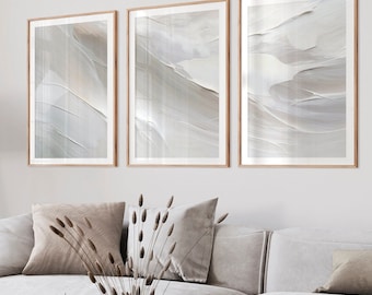 May include: Three framed wall art prints featuring abstract, textured brushstrokes in shades of white, beige, and soft gray. The artwork is displayed in light wood frames, arranged horizontally on a plain wall above a white sofa.