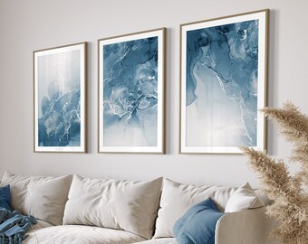 May include: A white sofa with blue throw pillows and a blue throw blanket in a living room with three framed abstract paintings with blue and white watercolour designs, a wooden coffee table, a floor lamp, and a vase with dried pampas grass.