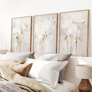 May include: Three framed abstract art prints with a white floral design on a light beige background. The prints are hanging on a white wall above a bed with beige and white bedding.