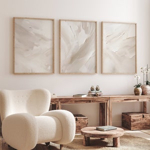 May include: A white armchair with a fluffy texture sits in front of a wooden console table with three abstract paintings above it. The console table has two wooden boxes on the bottom shelf and a small round wooden table with a book on top in front of it.