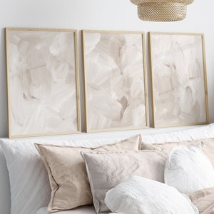 May include: Three framed abstract paintings with a beige background and white brushstrokes hang above a bed with white and beige linen bedding.