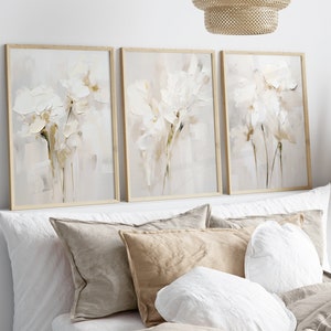 May include: Three framed abstract paintings with a white background and white flowers in shades of beige and brown.