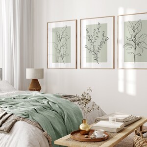 K&ouml;nnte beinhalten: Drei gerahmte Drucke von Strichzeichnungen von Pflanzen auf hellgr&uuml;nem Hintergrund. Die Drucke h&auml;ngen an einer wei&szlig;en Wand &uuml;ber einem Bett mit einer gr&uuml;nen Wolldecke.