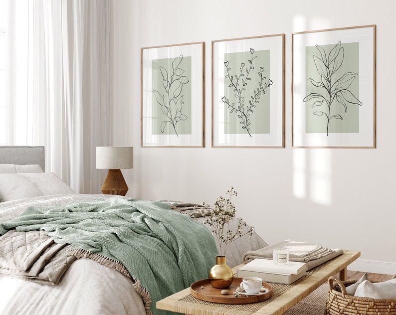 K&ouml;nnte beinhalten: Drei gerahmte Drucke von Strichzeichnungen von Pflanzen auf hellgr&uuml;nem Hintergrund. Die Drucke h&auml;ngen an einer wei&szlig;en Wand &uuml;ber einem Bett mit einer gr&uuml;nen Wolldecke.
