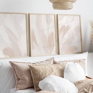 May include: Three framed abstract art prints in a light pink and white colour scheme. The prints are hanging above a bed with white and beige pillows.