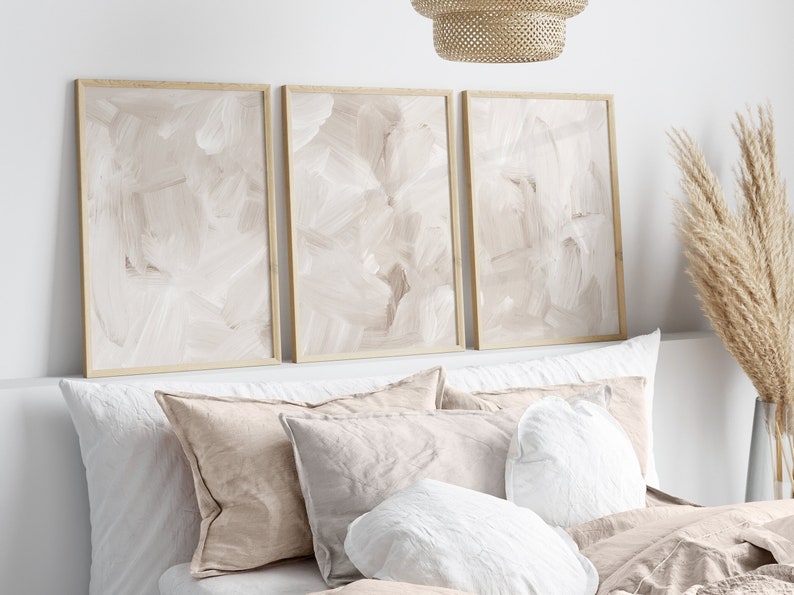 May include: Three framed abstract paintings with a beige background and white brushstrokes hang above a bed with white and beige linen bedding.