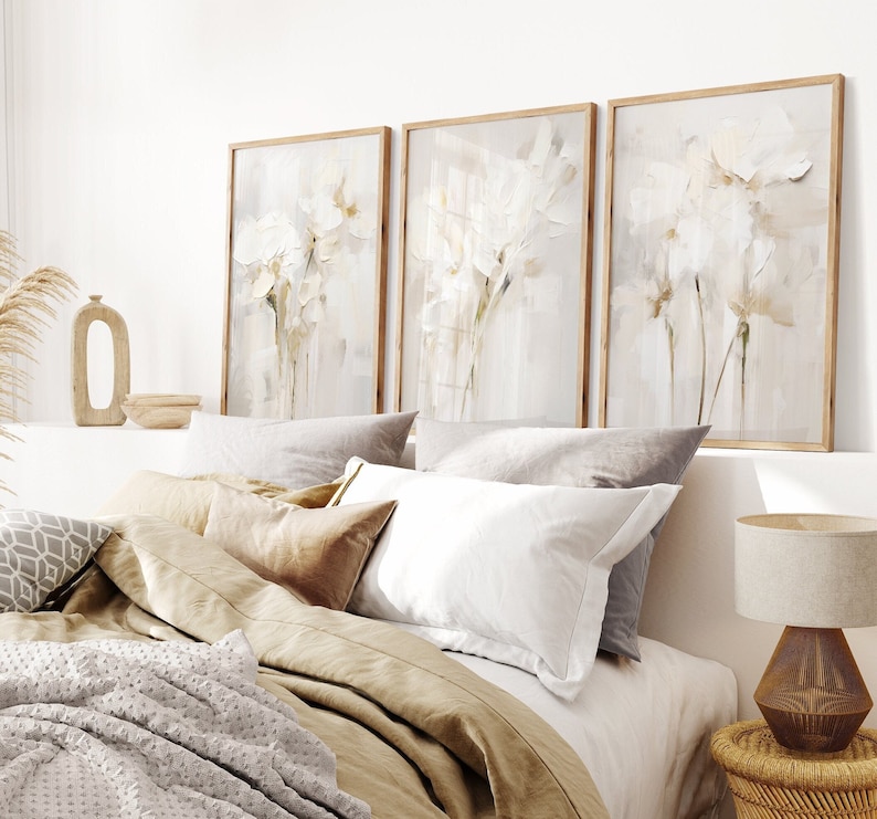 May include: Three framed abstract art prints with a white floral design on a light beige background. The prints are hanging on a white wall above a bed with beige and white bedding.