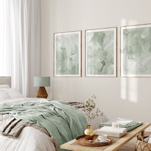 May include: A bedroom interior with a bed, a nightstand, a lamp, and three framed abstract art prints with a green and white color scheme. The prints are hung on a light beige wall. The bed has a white duvet, a green throw blanket, and a beige throw pillow. The nightstand is made of wood and has a lamp with a green shade. The floor is made of wood.