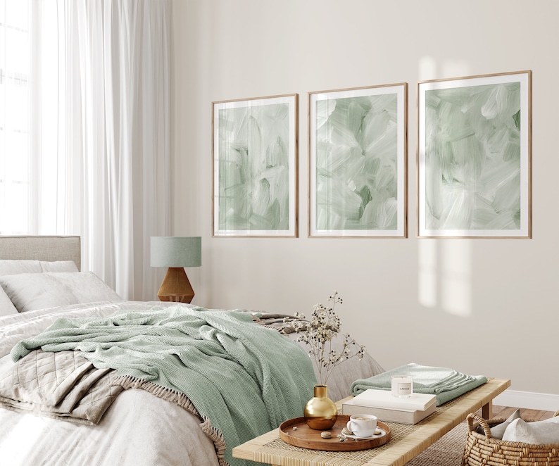 May include: A bedroom interior with a bed, a nightstand, a lamp, and three framed abstract art prints with a green and white color scheme. The prints are hung on a light beige wall. The bed has a white duvet, a green throw blanket, and a beige throw pillow. The nightstand is made of wood and has a lamp with a green shade. The floor is made of wood.