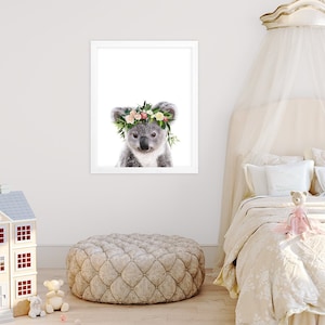 May include: A cute koala wearing a flower crown with pink, white, and green flowers. The koala is looking at the camera with a sweet expression. The background is white.