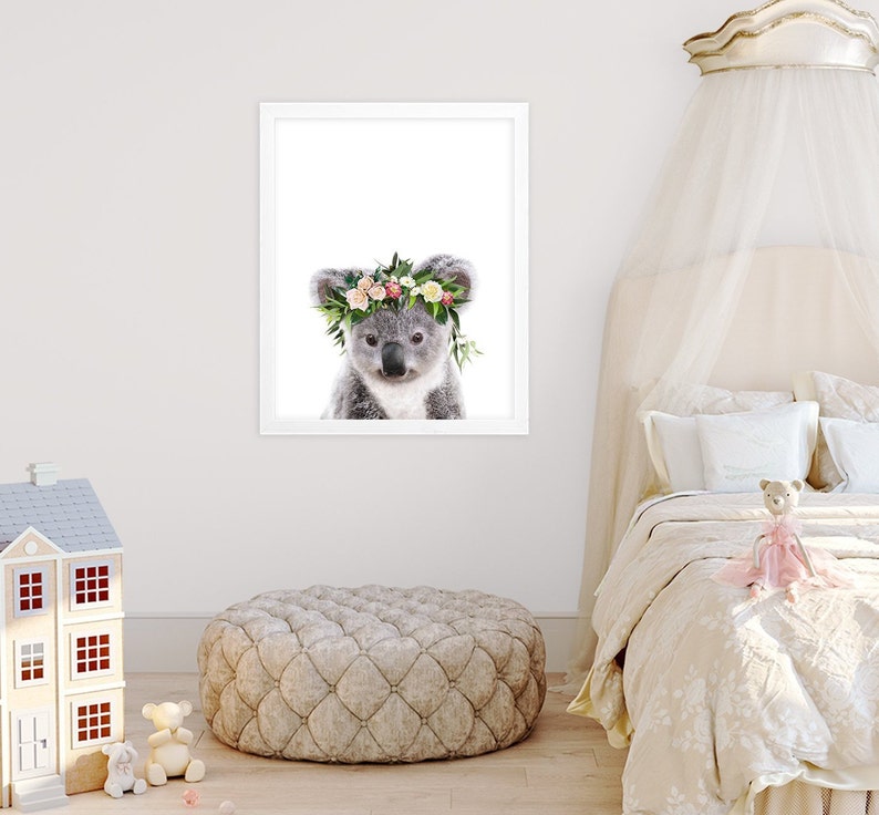 May include: A cute koala wearing a flower crown with pink, white, and green flowers. The koala is looking at the camera with a sweet expression. The background is white.