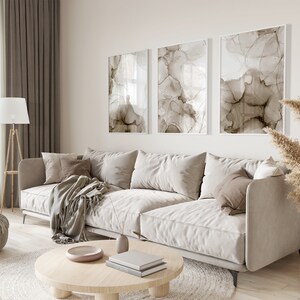 Puede incluir: Un sof&aacute; beige claro con una manta y almohadas en una sala de estar con una mesa de caf&eacute;, una l&aacute;mpara de pie y un jarr&oacute;n con hierba de la pampa seca. Tres pinturas abstractas con tonos marrones y blancos cuelgan de la pared.