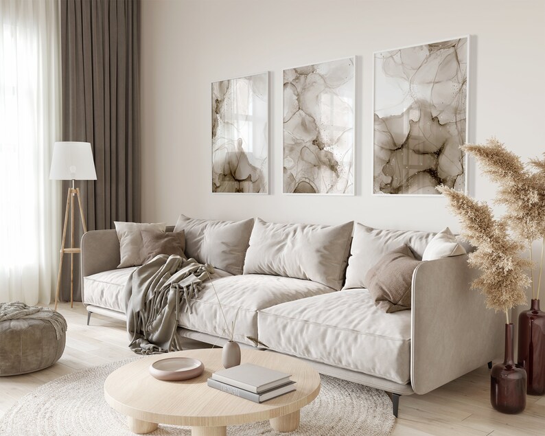 Puede incluir: Un sof&aacute; beige claro con una manta y almohadas en una sala de estar con una mesa de caf&eacute;, una l&aacute;mpara de pie y un jarr&oacute;n con hierba de la pampa seca. Tres pinturas abstractas con tonos marrones y blancos cuelgan de la pared.