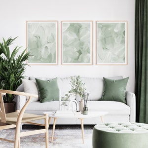 May include: Three abstract green paintings with white brushstrokes in wooden frames hang above a white sofa with green throw pillows. A potted plant, a vase with greenery, and a diffuser are on a white coffee table in front of the sofa.