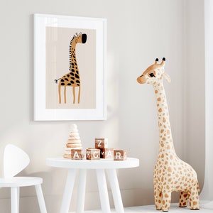 May include: A white framed print of a cartoon giraffe with brown and black spots on a light brown background. A stuffed giraffe toy with brown and white spots stands on a white floor. A white table with a wooden stacking toy and wooden alphabet blocks sits in front of the print. A white chair with bunny ears sits next to the table.