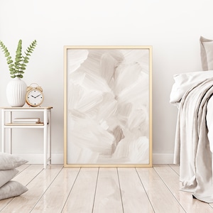 May include: A white bedroom with a bed, a bedside table, and a framed abstract painting. The bedside table has a white vase with green leaves, a clock, and a book. The bed has white bedding and a throw blanket.