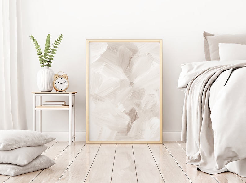 May include: A white bedroom with a bed, a bedside table, and a framed abstract painting. The bedside table has a white vase with green leaves, a clock, and a book. The bed has white bedding and a throw blanket.