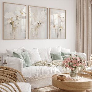 May include: Three framed abstract floral prints in muted tones of white and beige hang on a white wall above a white sofa with light green throw pillows. A vase of pink flowers sits on a wooden coffee table.